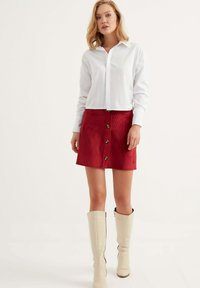 Camicia bianca a bottoni abbinata a una gonna in velluto a coste rossa con bottoni frontali e orlo svasato, completata da stivali alti fino al ginocchio color crema.