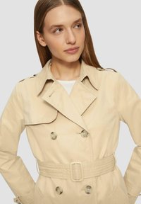 Beige trenchcoat met dubbele knopen, een ceintuur in de taille en schouderepauletten. Beschikt over een klassieke kraag en een gladde stoftextuur.