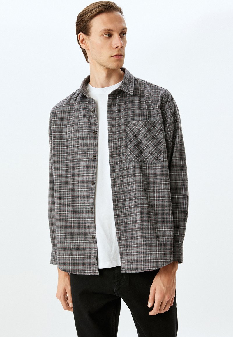 Camisa de cuadros gris con diseño abotonado, que presenta un bolsillo en el pecho y mangas largas. Lleva una camisa blanca debajo y se combina con pantalones negros.