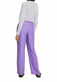 Osoba w wysokich, szerokich spodniach w kolorze lawendowym, białym dzianinowym swetrze do brzucha i czarnych butach, pokazana od tyłu.