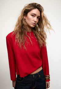 Maglione rosso corto con maniche lunghe e scollo a coste, abbinato a jeans scuri. La modella ha capelli mossi e indossa un braccialetto a un polso.