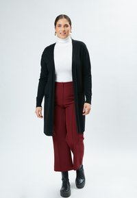 Zwarte lange cardigan met zakken over een witte turtleneck, gecombineerd met bordeaux brede broek en zwarte plateau laarzen.