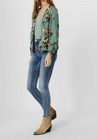 Blomstret jakke i grøn med puffede ærmer, lagdelt over en lysegrøn top. Blå skinny jeans og beige ankelstøvler fuldender looket.