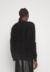 Eine Frau mit geflochtenem Haar trägt einen schwarzen strukturierten Pullover, goldene Creolen und einen schwarzen Lederrock. Sie wird von hinten vor einem schlichten weißen Hintergrund gezeigt.