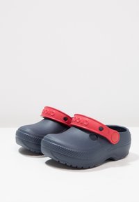 Granatowe drewniaki z czerwonym paskiem, wykonane z elastycznego materiału. Posiadają otwory wentylacyjne i teksturowane podeszwy zapewniające przyczepność. Lekka konstrukcja.