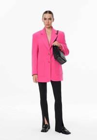 Vaaleanpunainen ylisuuressa bleiserissä on kaksi etupocketia, ja se on yhdistetty mustiin slim fit -housuihin, joissa on sivuaukot, sekä mustiin loafereihin.