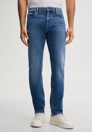 Man die een blauwe slim-fit spijkerbroek en witte sneakers draagt, staande tegen een effen achtergrond.