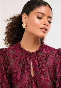 Bordová priesvitná blúzka s kvetinovou výšivkou, volánikovým výstrihom a detailom s kľúčovou dierkou. Zlaté výrazné náušnice so skrúteným dizajnom.