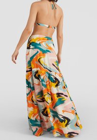 Mujer con una larga falda maxi de un patrón abstracto y colorido y un bikini halter a juego, de espaldas frente a un fondo liso.
