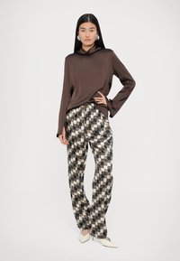Top lung cu maneci lungi din satin maro, cu guler tip cowl, asortat cu pantaloni lejeri cu model în carouri alb-negru și pantofi albi cu vârf ascuțit.