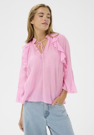 Femme portant un chemisier rose ample avec des volants et un col à nouer, associé à un jean taille haute bleu clair, main dans la poche.