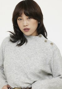 Hellgrauer Pullover mit hohem Kragen, gerippter Textur und drei dekorativen Knöpfen an der linken Schulter. Verfügt über ein dezentes diagonales Muster.