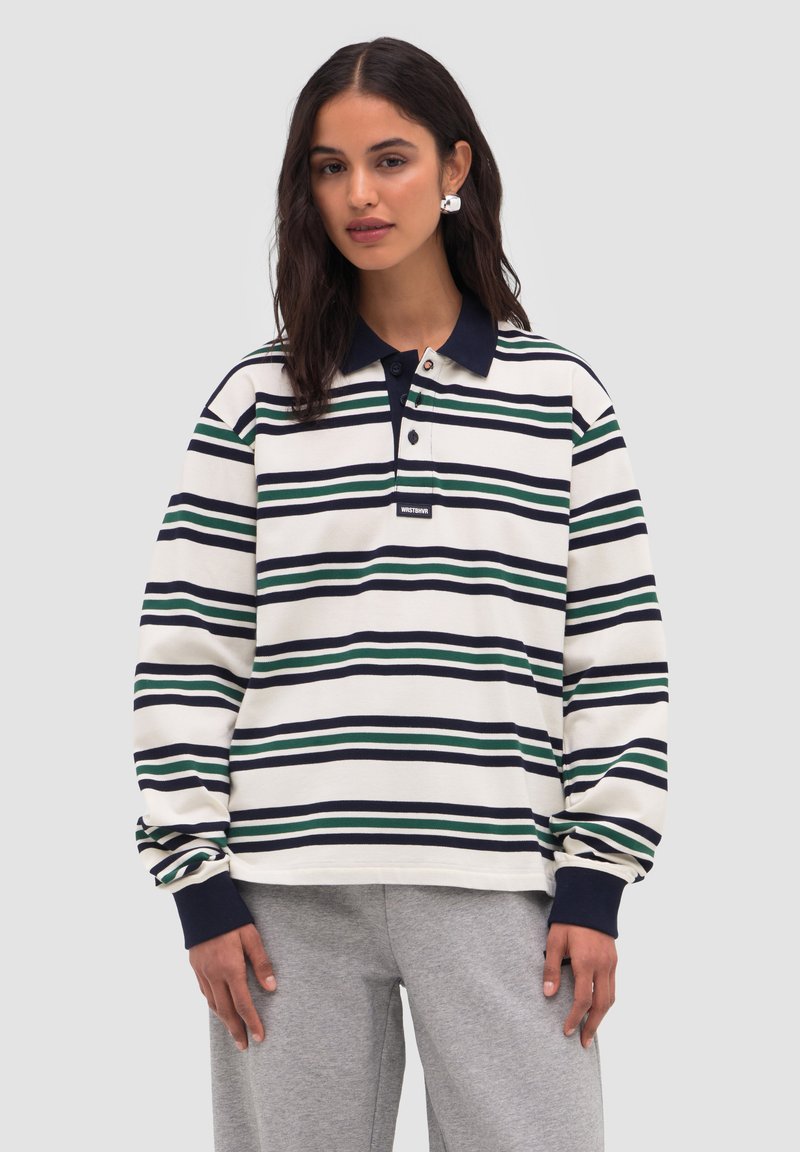 Nuori nainen, jolla on pitkät tummat hiukset ja valkoinen poolopaita, jossa on laivastonsiniset ja vihreät raidat, sekä harmaat vetoketjuhousut, seisoo yksinkertaisen taustan edessä.