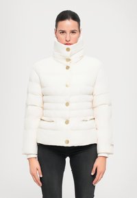 Giacca a collo alto bianca in stile puffa, con bottoni oro, tasche con zip e design trapuntato. Abbinata a pantaloni neri.
