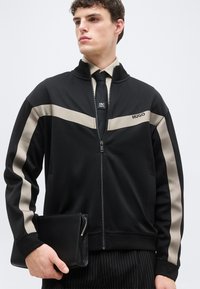 Νέος άνδρας που φοράει μαύρο μπουφάν με φερμουάρ και μπεζ ρίγες, μαύρη γραβάτα με λογότυπο, παντελόνι με λεπτές ρίγες, κρατώντας μια μαύρη τσάντα με φερμουάρ.