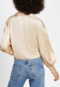 Satinbeige blus med kragenek, ballongärmar och knäppte manschetter, tillsammans med blå högmidjade denimjeans.