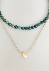 Zwei geschichtete Halsketten: Oben mit facettierten grünen Perlen, unten mit einer goldenen Kette und einem großen, cremefarbenen Perlenanhänger.