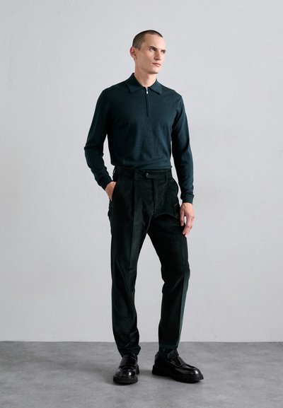 Moški v temno zeleni dolgem zip polo majici, visokih plisiranih črnih hlačah in črnih loaferjih stoji z rokami v žepih ob sivi steni.