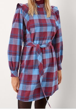 Femme portant une robe à carreaux ceinturée avec des épaules bouffantes dans des tons bordeaux, bleu et violet, associée à des collants noirs.
