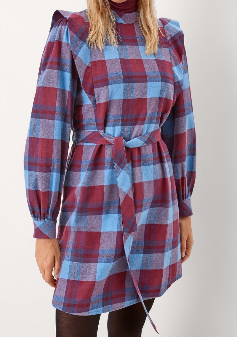 Femme portant une robe à carreaux ceinturée avec des épaules bouffantes dans des tons bordeaux, bleu et violet, associée à des collants noirs.