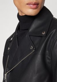 Zbliżenie na osobę w czarnej skórzanej kurtce z srebrnymi zamkami błyskawicznymi i guzikami, noszącą czarny sweter z golfem.