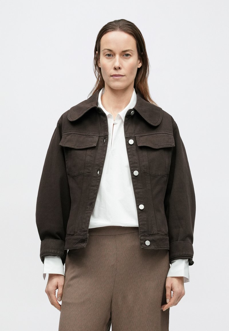Veste en denim marron avec un large col, des poches avant et des boutons-pression. Portée par-dessus une chemise blanche, associée à un pantalon beige.