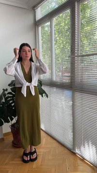Γυναίκα που διορθώνει τα μαλλιά της, φοράει ένα ελαιί πράσινο φόρεμα και λευκό μπλουζάκι με κόμπους, στέκεται κοντά σε ένα μεγάλο παράθυρο με περσίδες και μια γλάστρα.