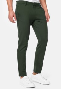 Slim-fit, dark green pants made from a smooth fabric, featuring a zip fly, front pockets, and subtle seam detailing. Paired with white sneakers.