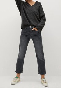 Persoon met een donkergrijze V-hals trui, hoog-gewijze rechte zwarte jeans en witte sneakers met beige en groene details.