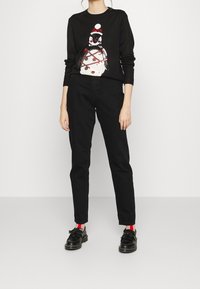 Pullover nero con design di pinguino e accenti rossi e bianchi, abbinato a pantaloni neri e scarpe nere con dettagli di calzini rossi.