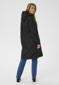 Schwarzer, gesteppter langer Mantel mit Kapuze, der ein diagonales Muster aufweist und Druckknöpfe am Saum hat. Kombiniert mit blauen geraden Jeans.