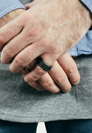 Mustaläbilise kuuepoolse metalli sõrmus heleda nahatooni käel, mis toetub teisele käele, mis on riietatud sinise ruudulise särkide ja halli kangaga.
