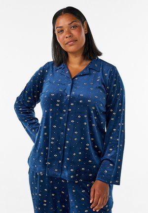Vrouw met schouderlang donker haar draagt blauw patroon lang mouw knoop pyjama top en bijpassende broek, staand met hand op heup.