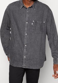 Camicia di denim grigia con colletto a bottoni, tasca sul petto e cuciture visibili. Caratterizzata da una vestibilità comoda e maniche lunghe.