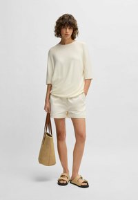 Light cream knit top, ivory shorts, woven beige bag, and tan sandals with decorative hardware. Model stands against a neutral background.