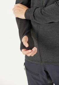 Giacca zip-up grigia scura con fori per il pollice nelle maniche, caratterizzata da un tessuto testurizzato e un design aderente, indossata sopra pantaloni scuri con tasche.