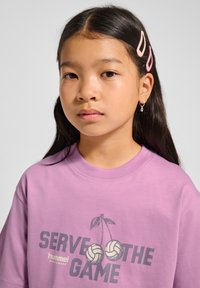 Lila T-Shirt mit kurzen Ärmeln, bedruckt mit einem Design aus Kirschen und Volleyballs, Schriftzug "SERVE THE GAME" und gemütlichem Schnitt.