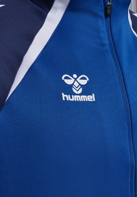 Veste de sport bleue avec fermeture éclair à l'avant et accents blancs. Elle a une texture lisse et affiche le logo "hummel" de manière proéminente.