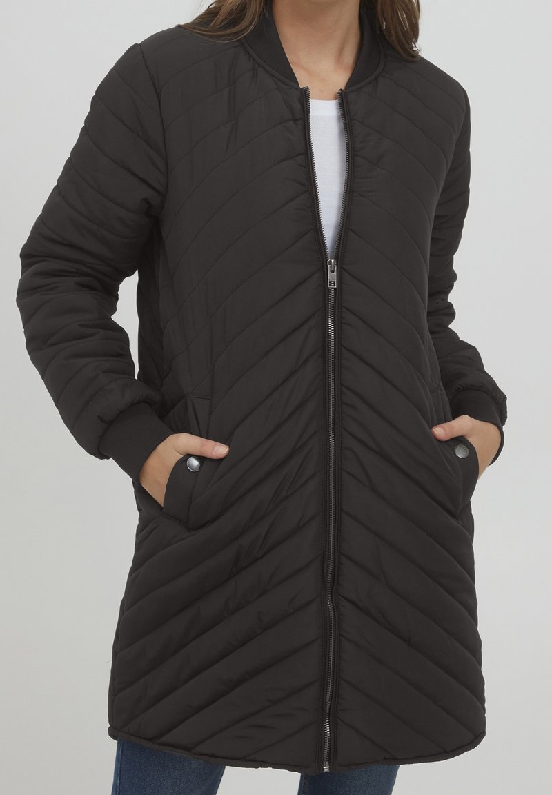 Femme portant une longue veste matelassée noire avec des coutures diagonales, une fermeture éclair à l'avant, des poches latérales et des poignets côtelés, par-dessus une chemise blanche et un jean.