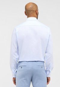 Camicia azzurra in tessuto liscio, con polsini abbottonati e colletto classico. Indossata con pantaloni su misura azzurri.