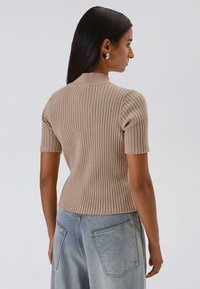 Una donna con capelli lunghi e diritti di colore scuro indossa un maglione beige a costine con maniche corte e jeans a vita alta di colore blu chiaro, voltata di spalle su uno sfondo neutro.