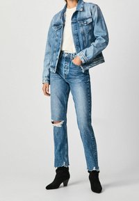 Osoba v světle modré džínové bundě a roztrhaných džínech, s bílým topem a černými kotníkovými botami na podpatku, stojící na jednotném pozadí.