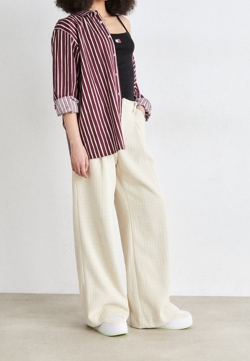 Camicia a righe bordeaux e bianca, canotta nera, pantaloni larghi in crema a coste, e sneakers bianche con dettagli verdi.