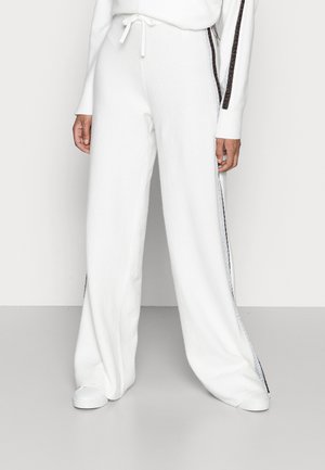 Persoon die losse witte broek met zwarte zijstrepen en witte sneakers draagt, staand tegen een effen achtergrond.