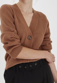 Cardigan marrone a coste con scollo a V profondo, chiusura con un bottone e maniche arrotolate, abbinato a pantaloni neri a vita alta con cintura.