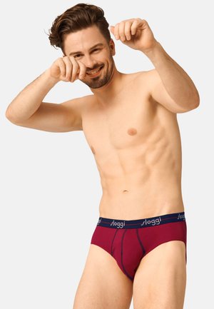 Smilende, barbrystet mand iført bordeaux Sloggi-trusser poserer med hævede hænder mod en ensfarvet lys baggrund.
