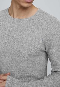 Pull en tricot gris clair avec un col rond, des manches longues et une poche plaquée sur la poitrine gauche avec une petite étiquette de marque.