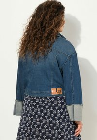 Giubbotto di jeans di colore blu scuro, con un design cropped, cuciture rifinite e un'etichetta in pelle marrone sul retro. Tessuto morbido e texturizzato.