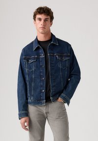 Veste en denim bleu foncé au design classique, avec des fermetures à boutons, deux poches avant et des coutures contrastantes. Portée avec une chemise noire.