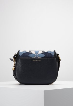 Sac bandoulière en cuir noir avec une fermeture dorée, logo Michael Kors et détail en daim bleu fleuri sur le dessus, exposé sur un socle blanc.